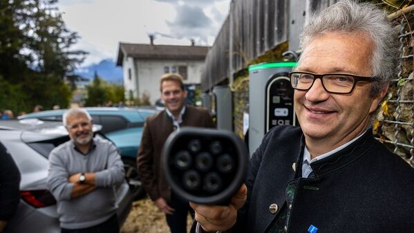 Ein Mann hält ein Ladegerät für Elektrofahrzeuge in der Hand, im Hintergrund sind zwei Personen und Autos zu sehen.