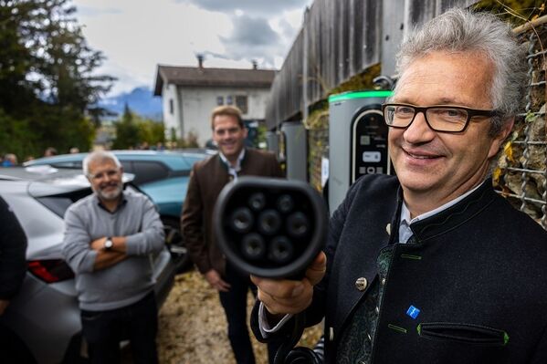 Ein Mann hält ein Ladegerät für Elektrofahrzeuge in der Hand, im Hintergrund sind zwei Personen und Autos zu sehen.