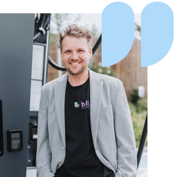 Ein Mann in einem grauen Blazer, der neben einer Maschine steht und lächelt, mit blauen Anführungszeichen.