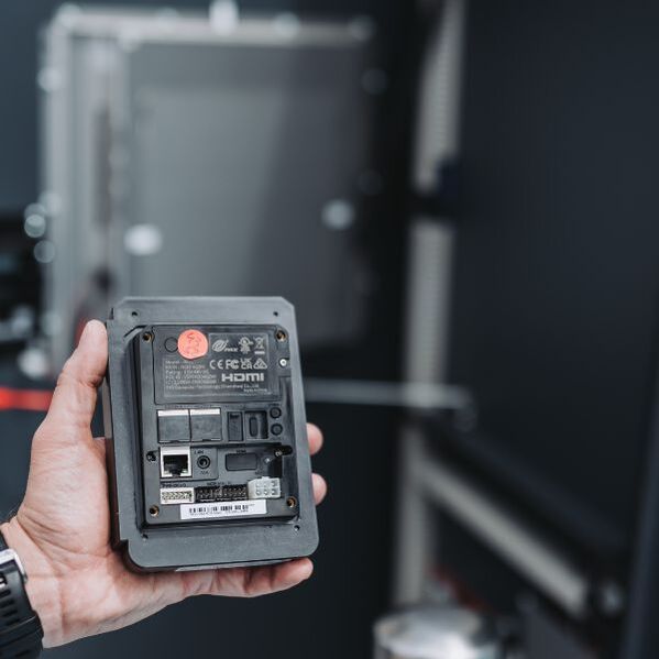 Eine Hand hält ein elektronisches Gerät, dessen Anschlüsse und Beschriftungen zu sehen sind; der Hintergrund ist unscharf.