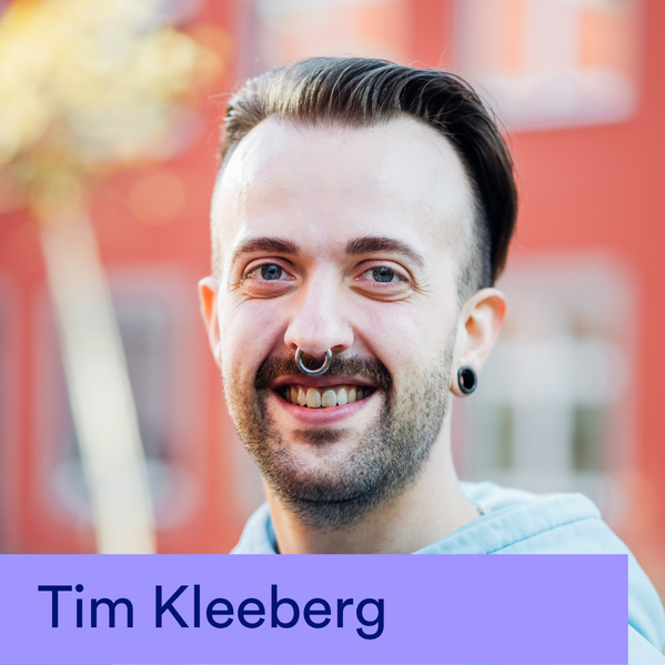 Eine lächelnde Person mit einem Bart und Piercings steht vor einem roten Gebäude.